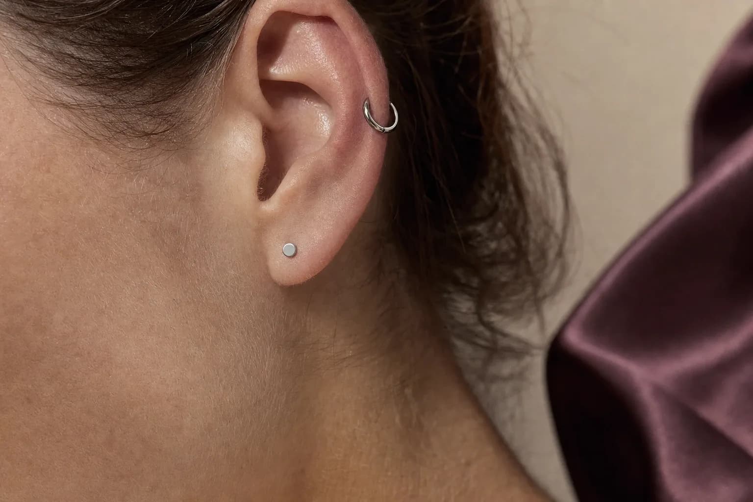 Ohr im Profil mit einem geschlossenen silberfarbenen Clicker im Helix und einem flachen Stecker im Ohrläppchen, dahinter unscharfe pflaumenfarbene Seide